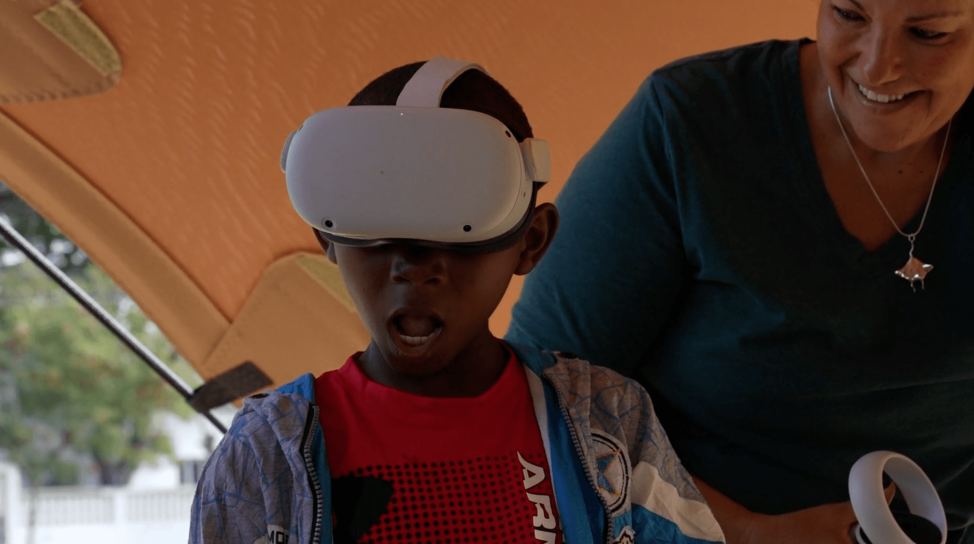 Child wearing a virtual reality headset with an open mouth, an adult smiling beside them.