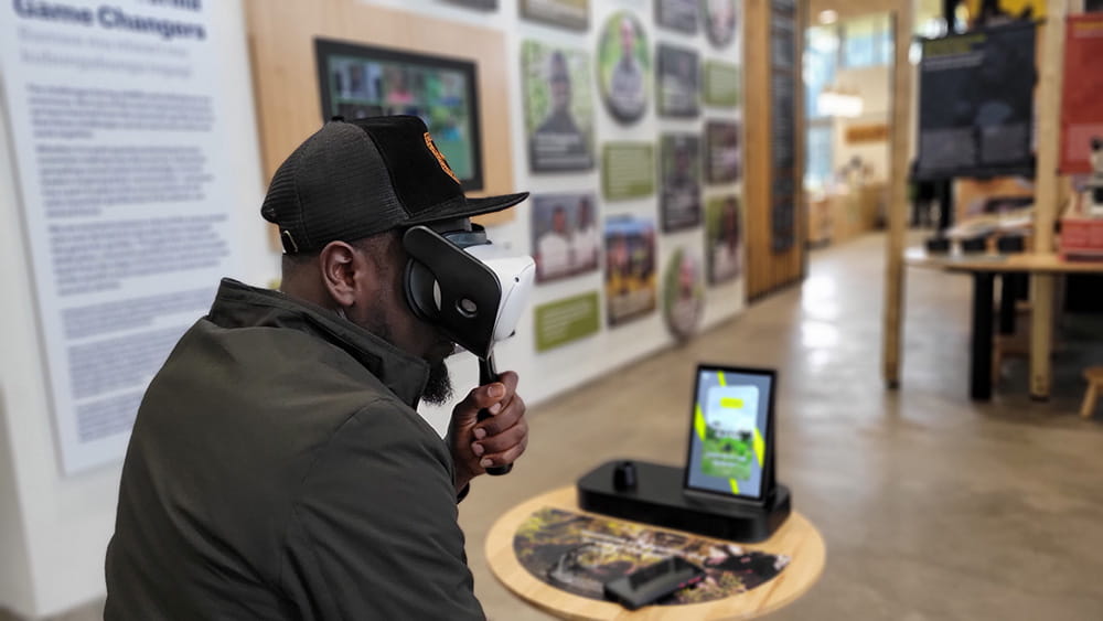 A visitor uses a custom-built VR headset and handheld controller at an interactive exhibition station, with interpretive displays visible in the background.